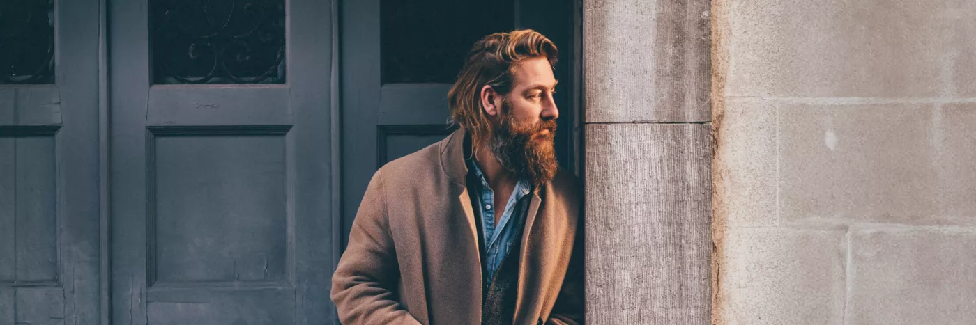 A man with brown hair and a long brown beard wearing a camel-colored coat over a denim shirt, leans on a door frame.