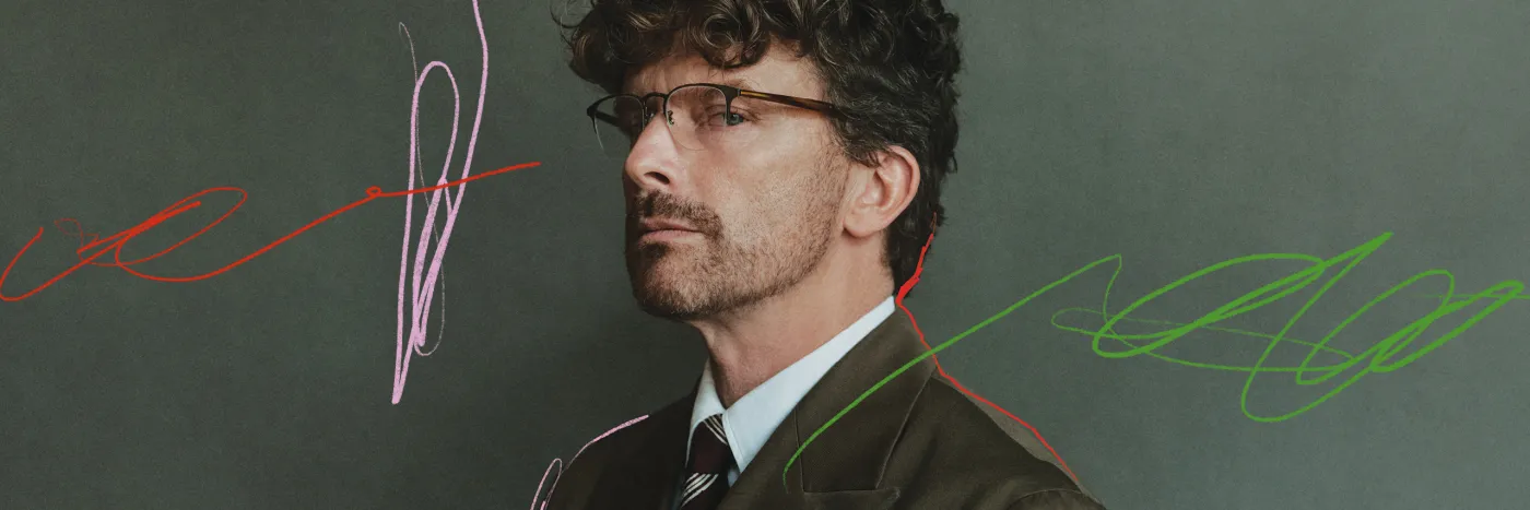 A man wearing glasses and a dark green suit looking sidelong against a grey-green background with subtle scribbled marks in pink, red and green.