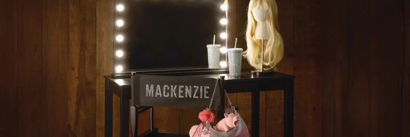 A long blonde wig and cup with a straw sit on a black dressing table with a mirror edged with lights and a folding chair with Mackenzie on the back and a pink handbag hanging off one side.
