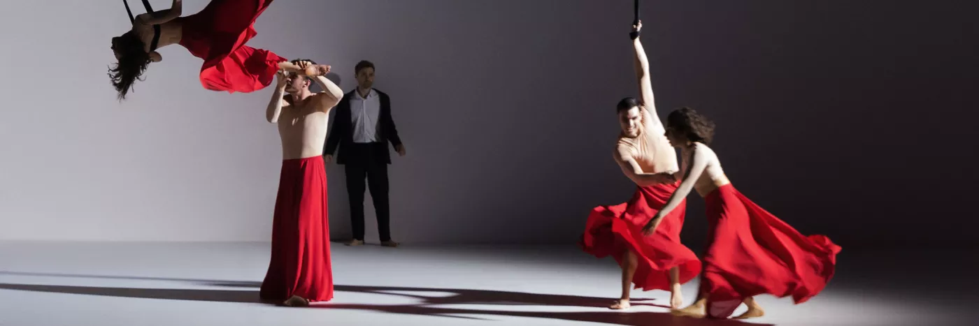 Two cast members wearing long red skirts, one is horizontal in the air attached to an aerial rope, the other assisting her movement