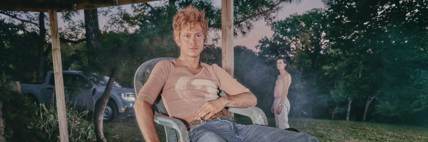 A white man wearing a beige top and baggy jeans louges in a plastic chair.