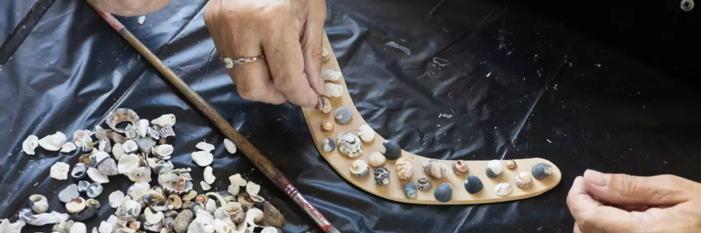 Two hands are placing small shells onto a light brown boomerang.