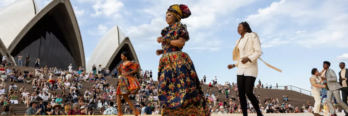 A group of people in colourful dress, walking in front of a crowd on the steps of the Sydney Opera House.