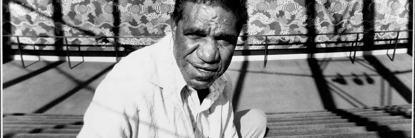 Jagamara photographed in black and white sitting on carpeted steps in front of the mural 'Possum Dreaming'.