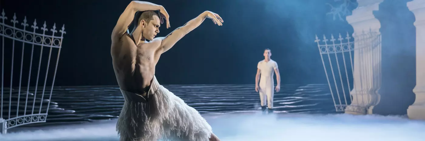 A topless man in a white feathery costume dancing under a set with a full moon on a blue lit stage.