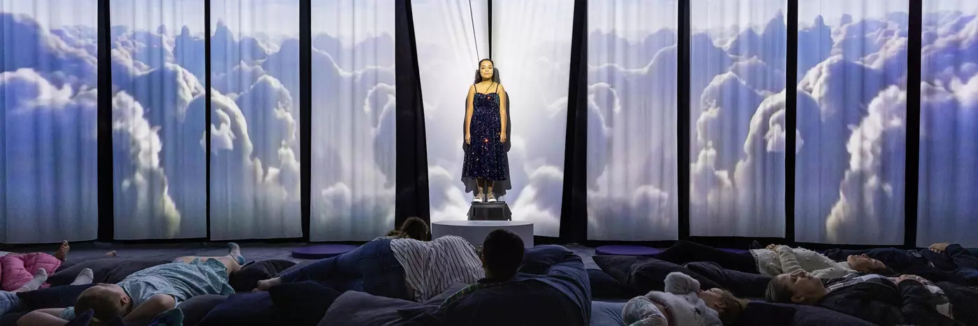 A young white woman with dark brown hair stands in front of a fabric background, where projections of clouds are being cast. An audience of meditation children and adults lie on pillows in front of her.