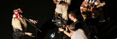 Three men filming a woman singing, whilst a man plays a guitar.
