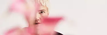 A person with short blonde hair standing behind some pink foliage against a white background.
