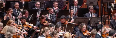An orchestra performing. We see men and women playing a variety of instruments including violins, oboes, trumpets and flutes.