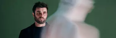 A white man with black hair and dark beard wears all black. He's in front of a green background and is looking at a shadow.