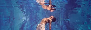 A shirtless man swimming underwater in a pool wearing a cloth skirt has his face looking up towards the sky,
