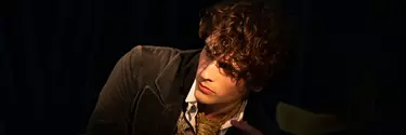 A man with light brown curly hair looks off into the distance in front of a black background. He wears a gold tie, white shirt and green jacket.