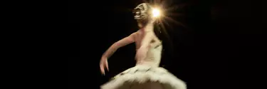 A ballerina wears a gold tutu and hairpiece. Her back is to the camera and her arms are stretched behind her, mid-dance move. A spotlight is seen behind her.