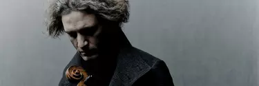 A 40-50 year old man with medium length grey hair wearing a coat looks down at the top of a string instrument.