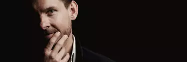 A close up of a white 30-40 year old man wearing a suit has his hand on his chin. Half his face is in darkness.