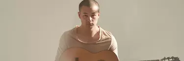 A young Asian man with shaved hair wears a beige top and holds an acoustic guitar.
