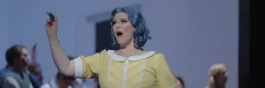 A woman in costume as a doll with a bobbed blue wig and yellow dress has her mouth open singing