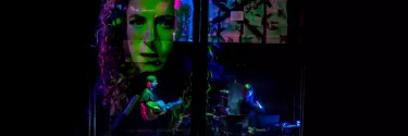 A man wearing a beanie plays the guitar while a woman next to him plays a keyboard, They are surrounded by a projection of a woman's face, and the lighting is green, purple and blue.