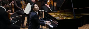 A Korean man wearing a black suit sits at a grand piano looking up and smiling