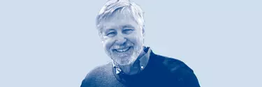 A white man with white hair and a beard smiling. He wears a blue sweater.