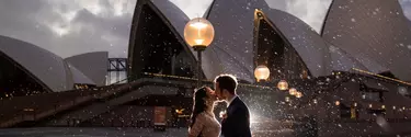 A woman in a wedding dress and a man in a suit kissing outside of the Sydney Opera House.