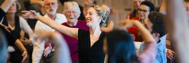 A woman in a black dress smiling, reaching out with an audience surrounding her.