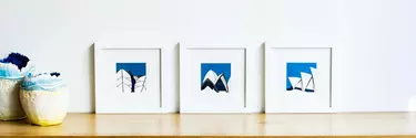 Three framed blue pictures resting on a wooden cabinet against a white wall.