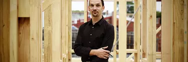 A man in a black shirt and long hair standing underneath wooden construction work.