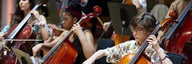 Two rows of young people seated in a room, playing cellos.