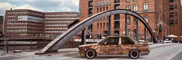 A BMW sedan covered in patterns of ochre, yellow, brown and blue parked on the pavement in front of high-rise buildings.