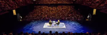 Two people sat on chairs on a stage in the middle of a full seated audience in a red lit theatre.