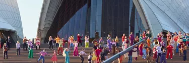 A large group of people wearing colourful clothes on the steps of the Sydney Opera House, viewed from a distance.