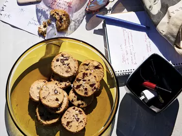 A yellow translucent glass plate with numerous salted butter and chocolate chunk shortbread on a tabletop. A notebook, pen, sheets of paper with writing and a container holding a charger and lipstick surround it.