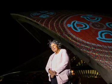 A woman in a white jacket against a textured colorful visual projected onto the Sydney Opera House's sails, smiling.