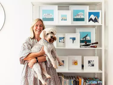 A woman holding a dog, standing in front of cabinet filled with framed pictures.