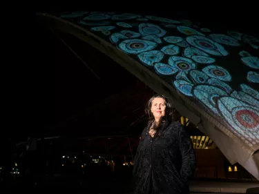 A woman in a black shirt against a textured colorful visual projected onto the Sydney Opera House's sails, smiling.