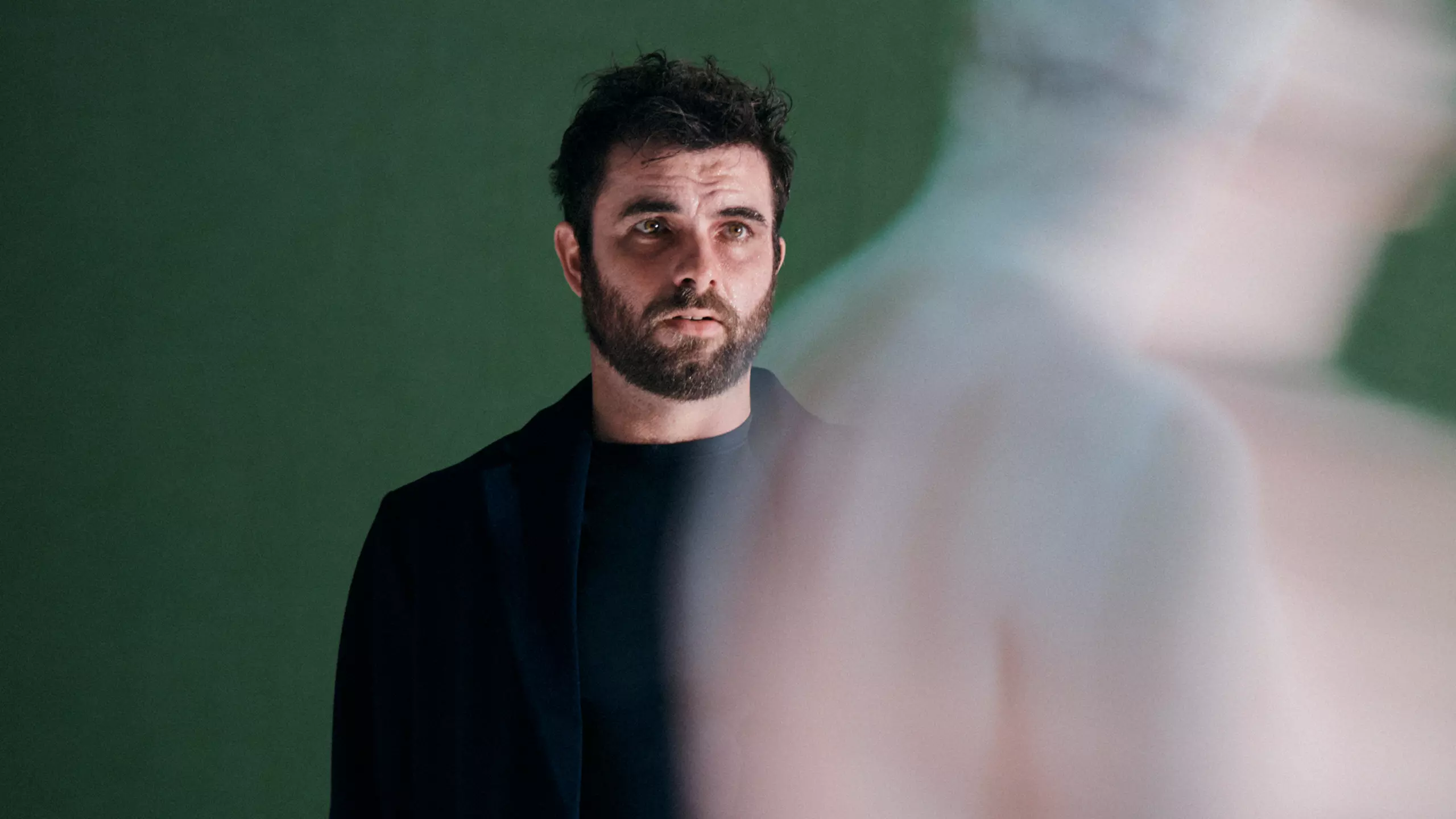 A white man with black hair and dark beard wears all black. He's in front of a green background and is looking at a shadow.