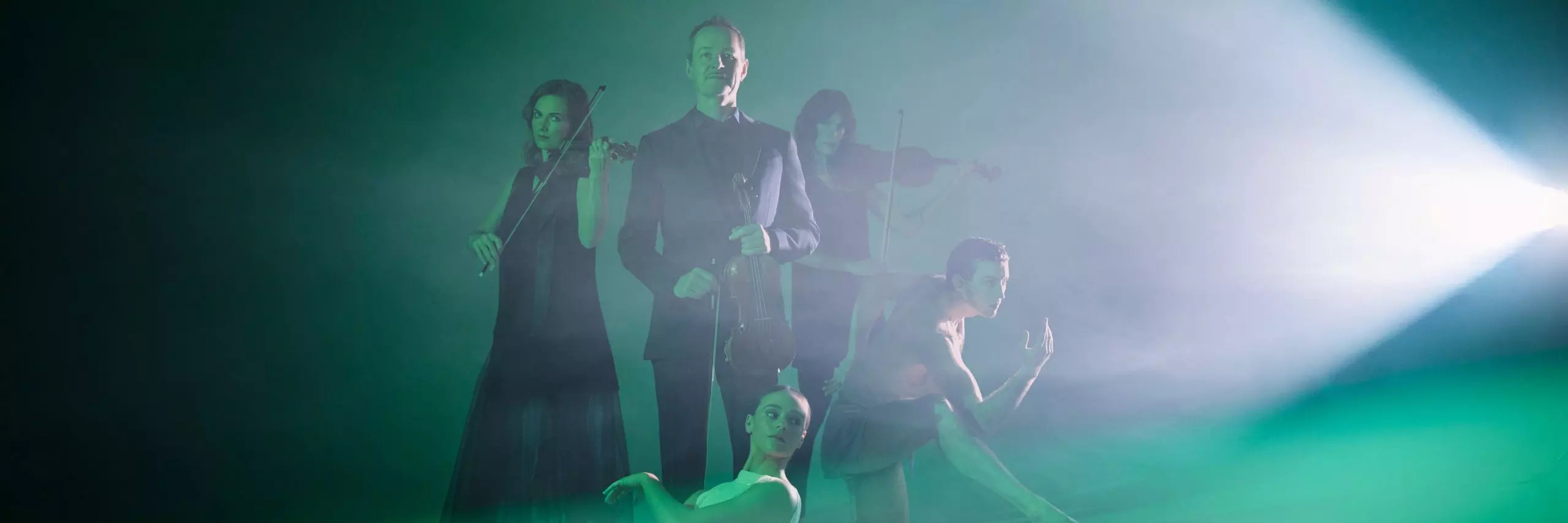 A white, 50-year old man holds a violin immersed in green light. Around him is two female violinists, a man dancer and a female ballerina.