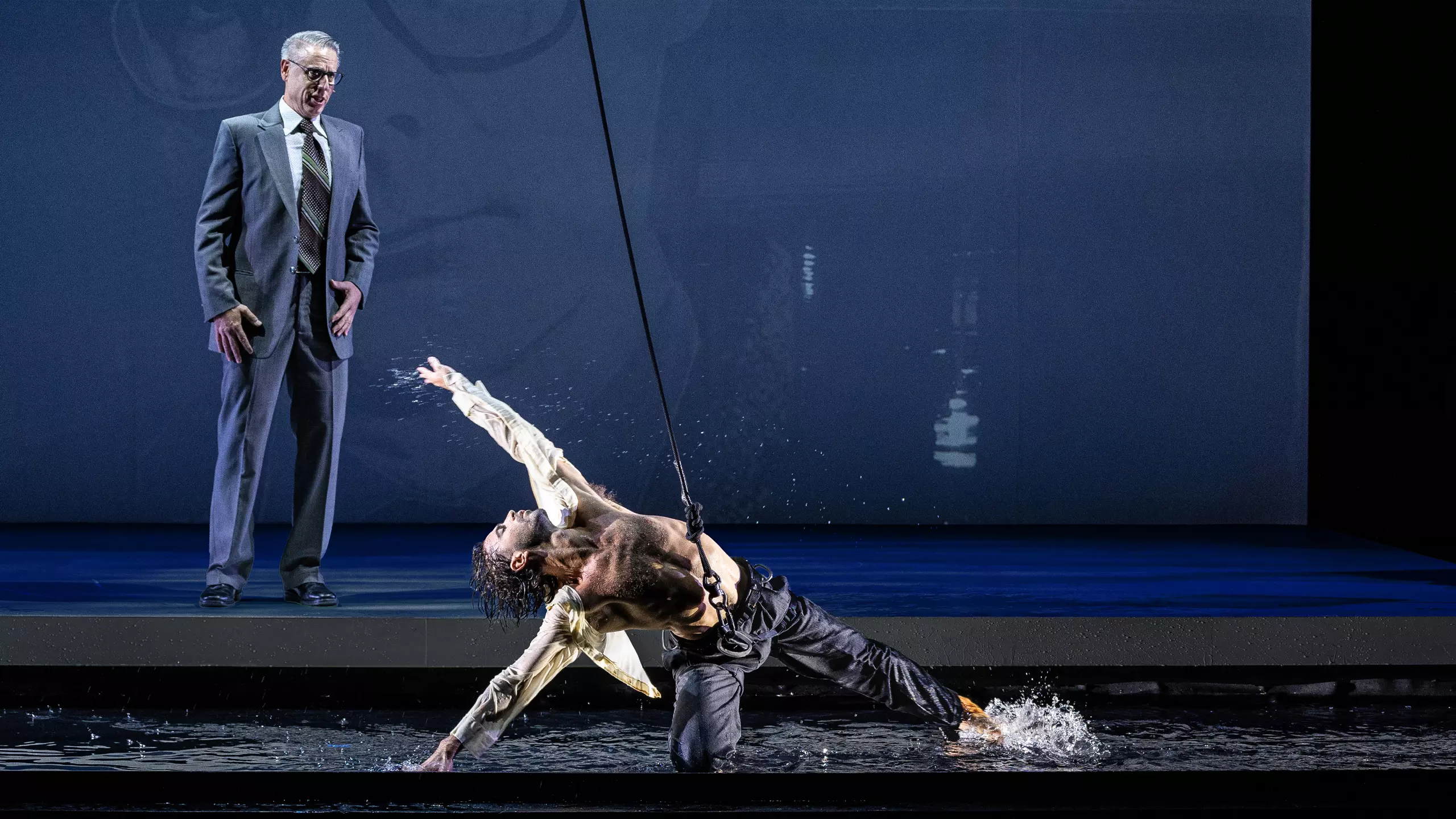 A white man with grey hair in a suit stands looking at a male dancer knelt on the floor. The dancer wears a harness and is dancing in water.