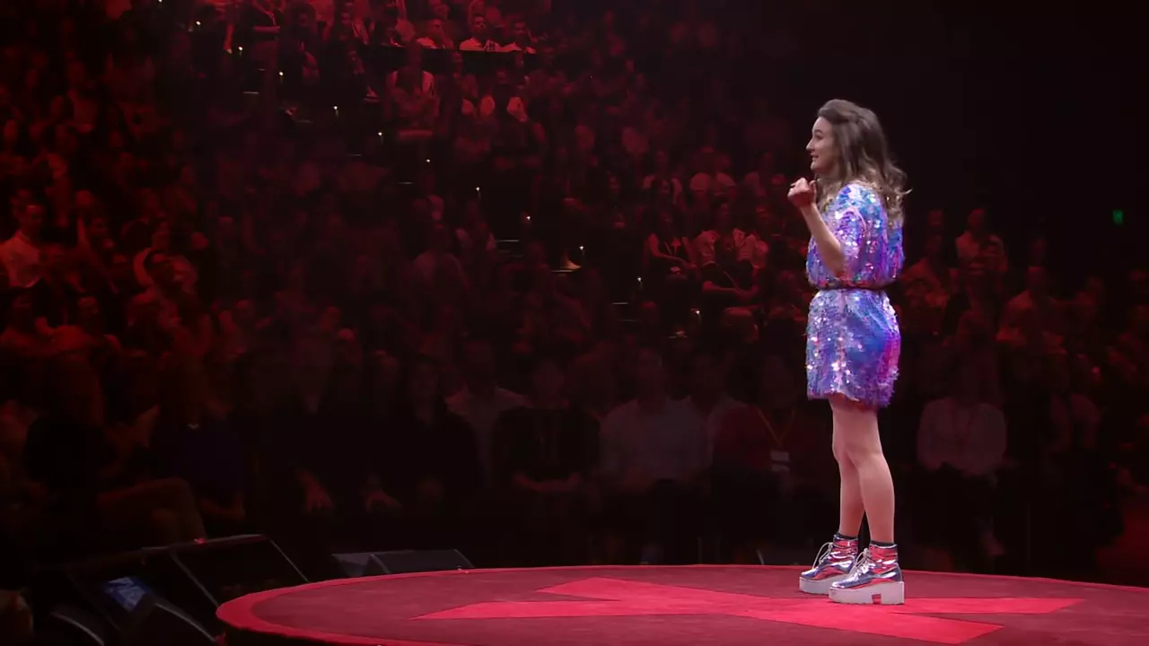 A woman in a purple sparkly dress and silver boots stood on a stage talking to an audience.