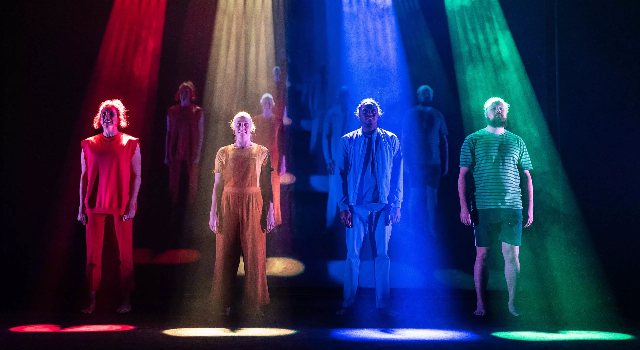 Four people wearing different prime colours. Their arms are out to the side, with finger splayed. They appear to be yelling. They are all stacked up behind one another and coloured lights are projected in the background.