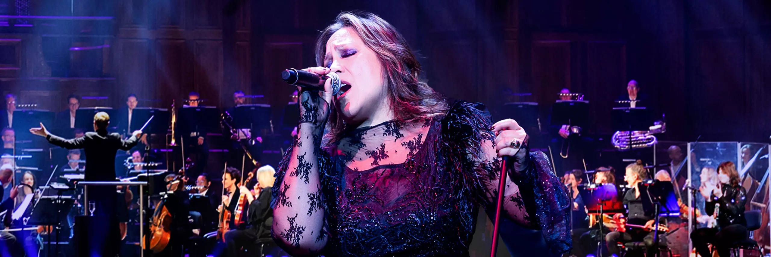 A Filipino-Australian woman with brown layered, shoulder-length hair holds a microphone in her right hand and the microphone stand in her left hand. She's singing on stage in front of an orchestra.