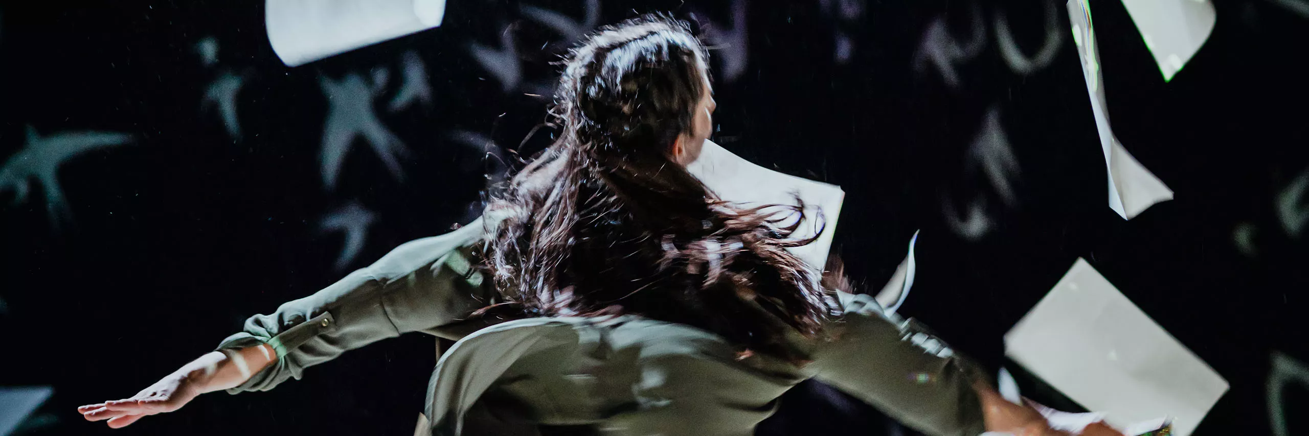 A women dances surrounded by falling paper. She is holding a book and her hair is in braids.