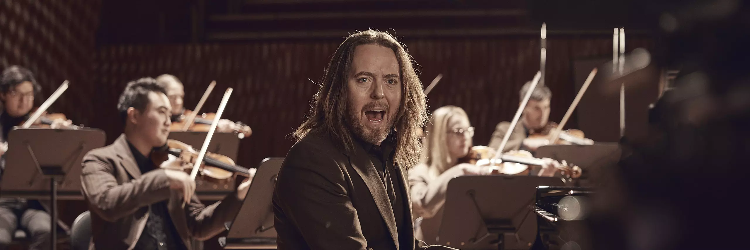 Tim Minchin signing and playing the piano, with violinist's playing in the background.